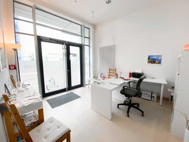 Praxis Eingang von innen, Bürotisch im Anmeldebereich mit Bürostuhl - Der Raum ist überwiegend in weißen Farben gestaltet. Im Vordergrund ist ein Holzstuhl zu sehen